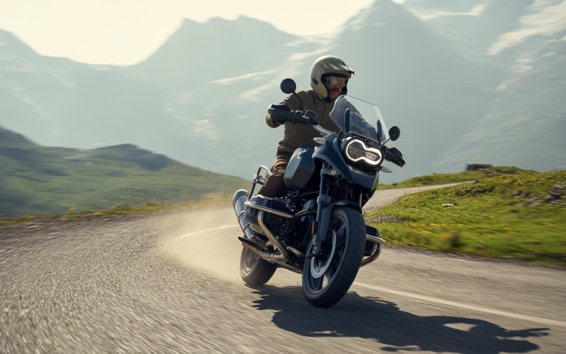 Un motard conduit une moto sur une route sinueuse, avec des montagnes en arrière-plan et de la poussière soulevée.