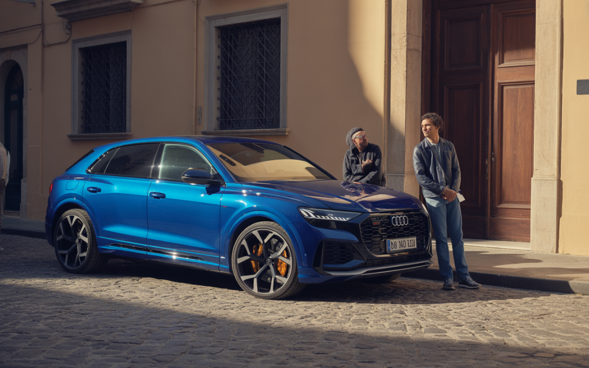 Une voiture Audi Q7 bleue stationnée dans une rue pavée, deux hommes discutent à proximité d’un bâtiment historique.