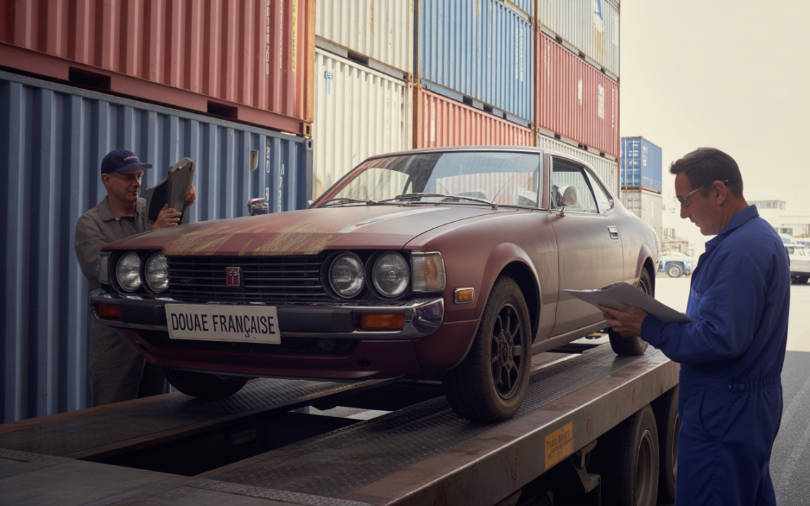 Deux hommes inspectent une voiture ancienne sur une remorque, entourés de conteneurs de transport. Étiquette "Douane Française" visible.