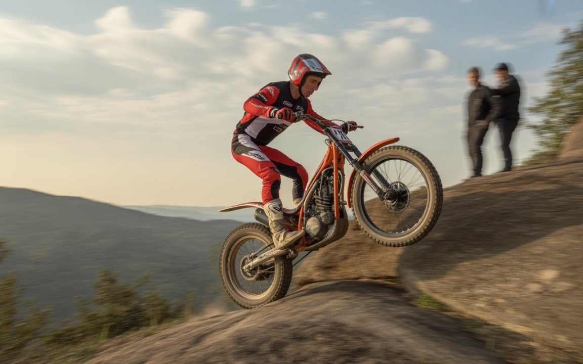 Un pilote de moto trial saute sur un rocher, avec des paysages montagneux à l’arrière-plan et deux spectateurs flous.