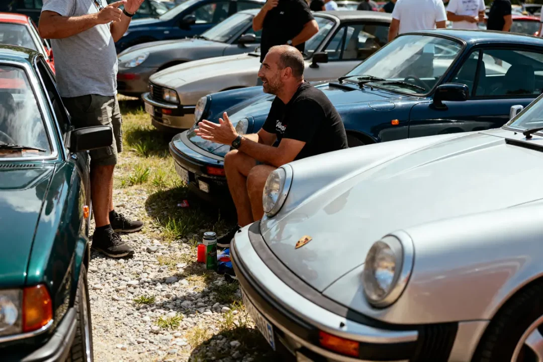Homme discutant avec d'autres, entouré de voitures de sport classiques dans un rassemblement en plein air, journée ensoleillée.