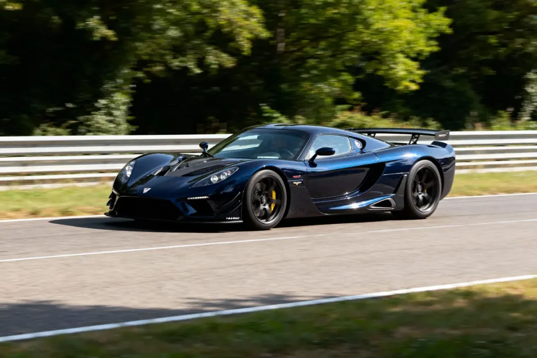 Voiture de sport noire roulant sur circuit bordé d'arbres et de glissières de sécurité.