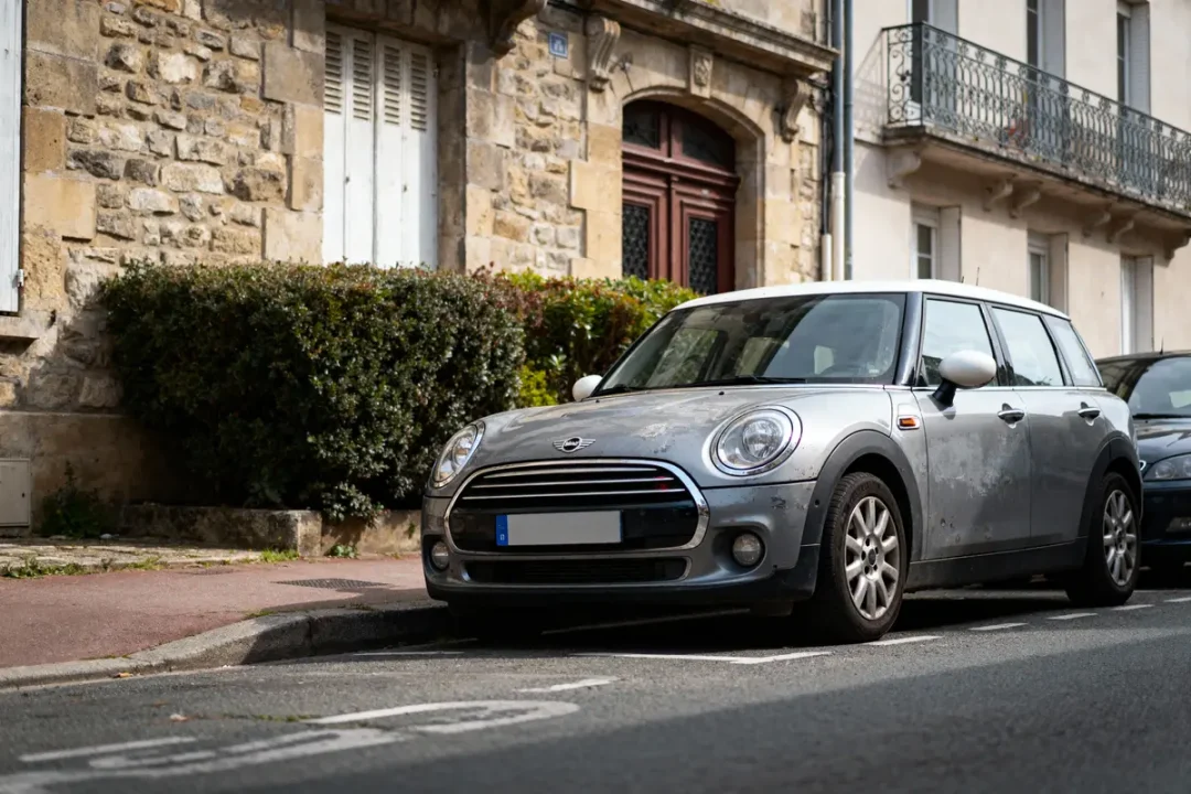 Voiture Mini Cooper grise garée devant bâtiment en pierre, arbustes à gauche, route et trottoir visibles.