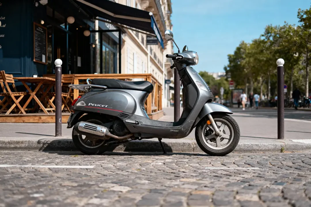 Scooter gris Peugeot stationné devant un café avec terrasse en ville, rue pavée, ciel bleu.