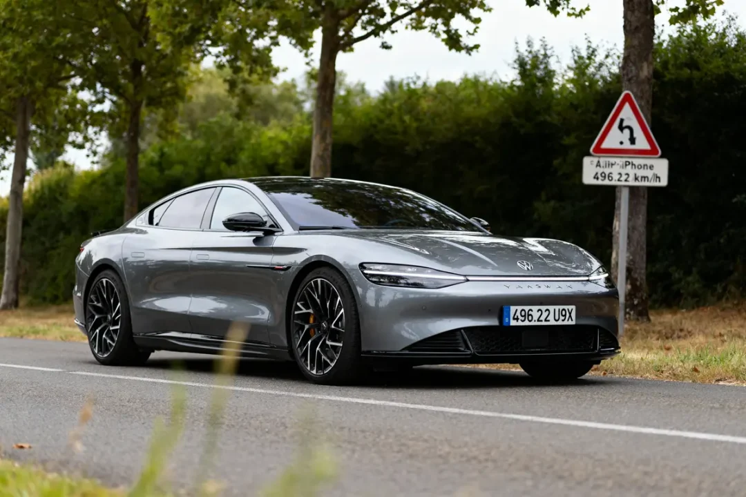 Voiture grise élégante stationnée, arbres en arrière-plan et panneau routier humoristique limitant la vitesse.