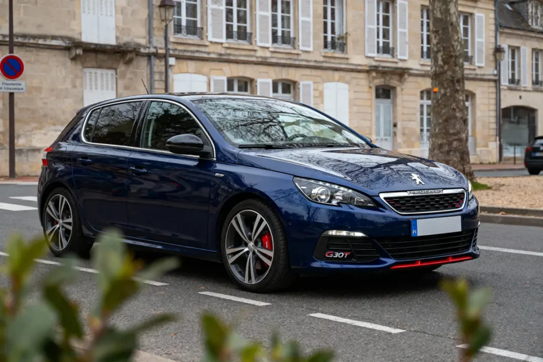Voiture Peugeot bleue stationnée devant bâtiment en pierre avec volets, rue bordée d'arbres.