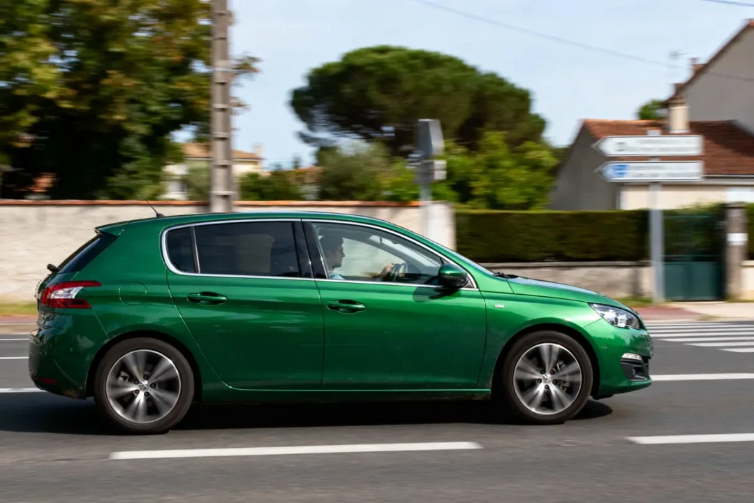Voiture verte en mouvement sur route, conducteur visible, arrière-plan urbain flou.