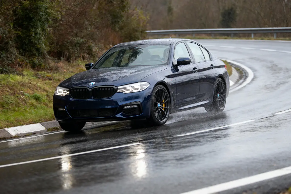 BMW bleue conduite sur route mouillée, virage, journée pluvieuse, arbres en arrière-plan.