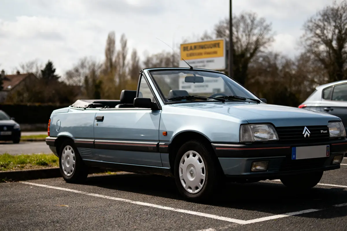 Voiture décapotable bleue Citroën garée sur un parking par temps nuageux. Arrière-plan arboré et signalisation.