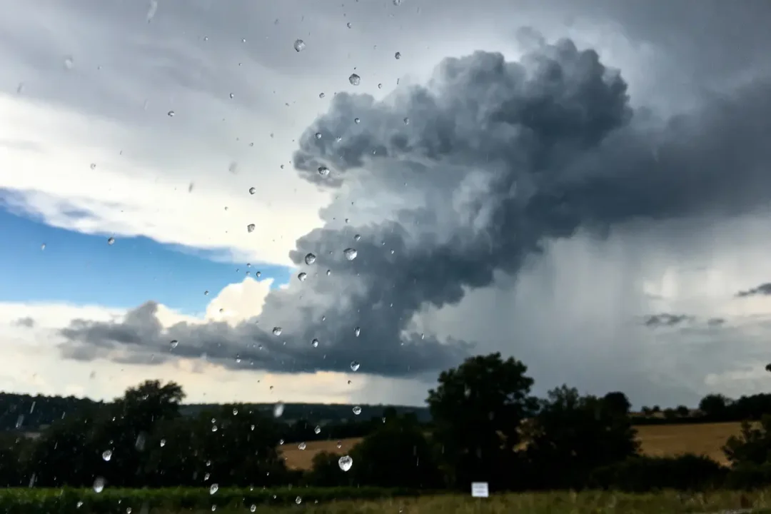 Gros nuage sombre menaçant, pluie sur fenêtre, ciel partiellement bleu. Paysage champêtre en arrière-plan.