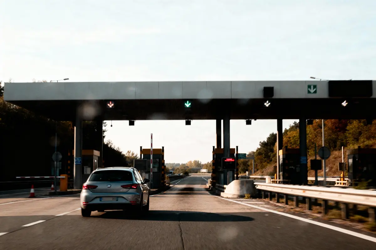 Voiture argentée passant un péage d'autoroute avec barrières et signalisations lumineuses.
