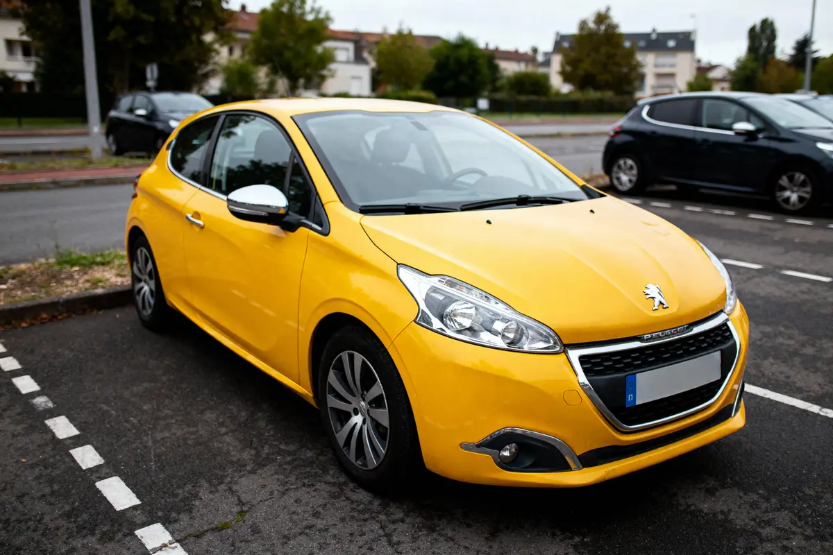 Voiture jaune Peugeot garée sur une place de parking, en environnement urbain avec bâtiments et végétation.