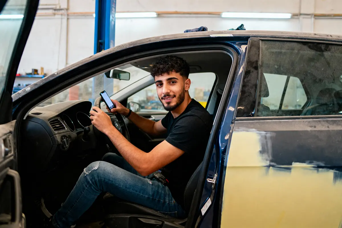 Jeune homme souriant dans une voiture, tenant un smartphone dans un atelier de réparation automobile.