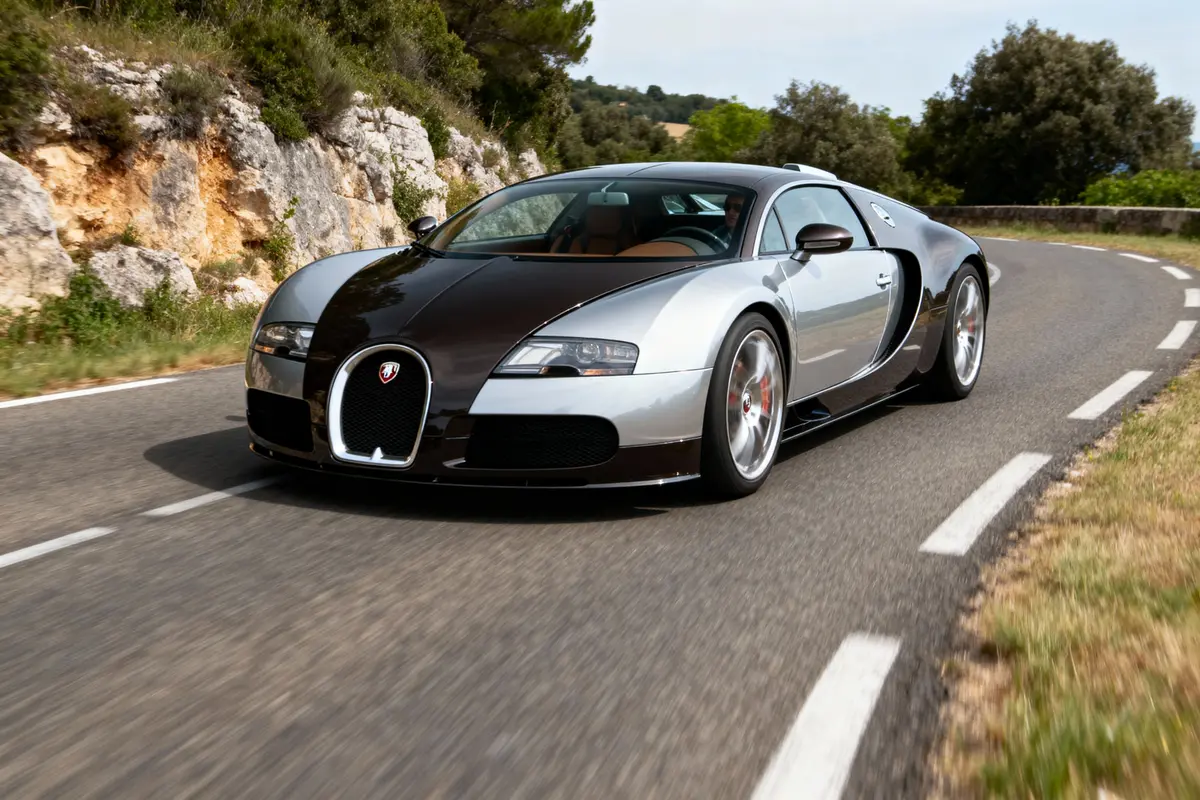 Voiture de sport grise et noire sur une route sinueuse, entourée de végétation et de rochers.
