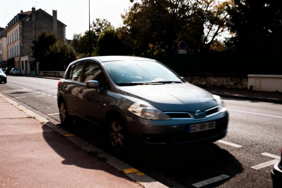 Voiture grise garée sur une rue ensoleillée bordée d'arbres et bâtiments résidentiels.