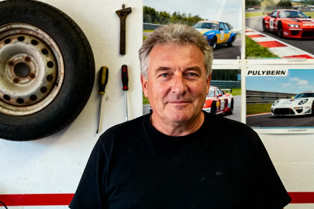 Homme souriant devant mur avec outils, pneu, et affiches de voitures de course en fond.