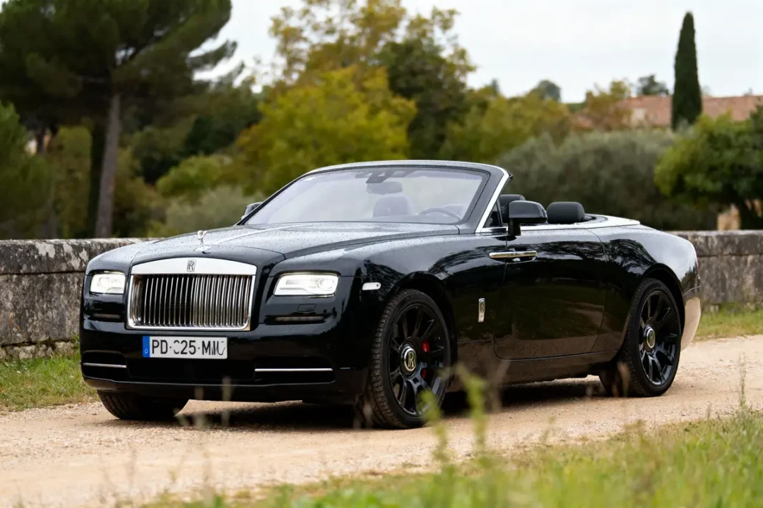 Voiture décapotable luxueuse noire sur chemin de terre, entourée de verdure, ciel nuageux.