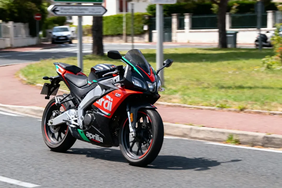 Moto Aprilia rouge et noire roulant sur route urbaine par temps ensoleillé, panneaux routiers en arrière-plan.