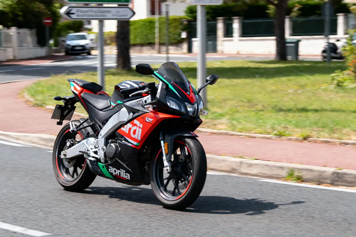 Moto Aprilia rouge et noire roulant sur route urbaine par temps ensoleillé, panneaux routiers en arrière-plan.