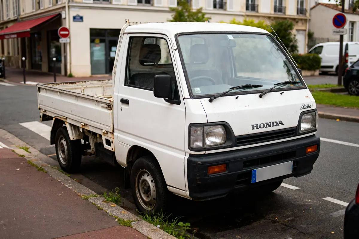 Petit camion utilitaire blanc Honda, garé dans une rue urbaine, devant un bâtiment, jour.