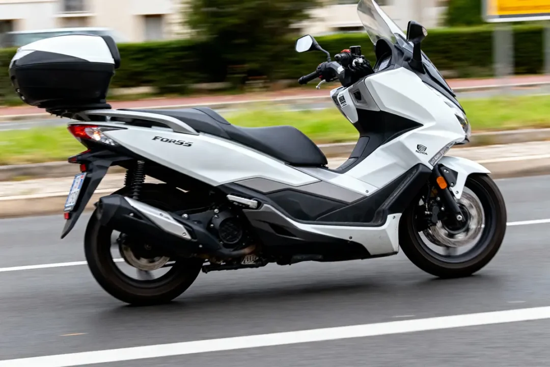 Scooter blanc Honda Forza 125 roulant sur la route avec top case noir, vue latérale.