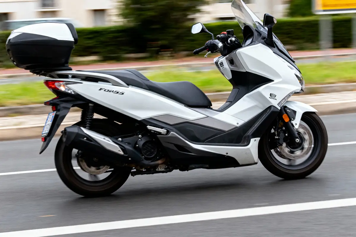 Scooter blanc Honda Forza 125 roulant sur la route avec top case noir, vue latérale.