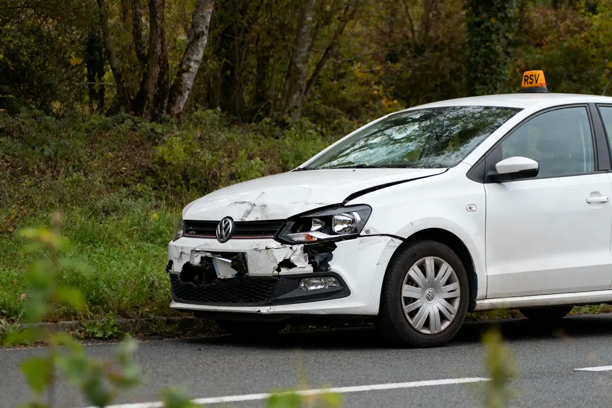 Voiture blanche accidentée sur le bord de la route, pare-chocs avant endommagé, en forêt.