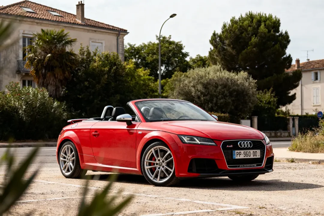 Voiture décapotable rouge Audi TT garée devant un bâtiment ancien et des arbres, ensoleillée.
