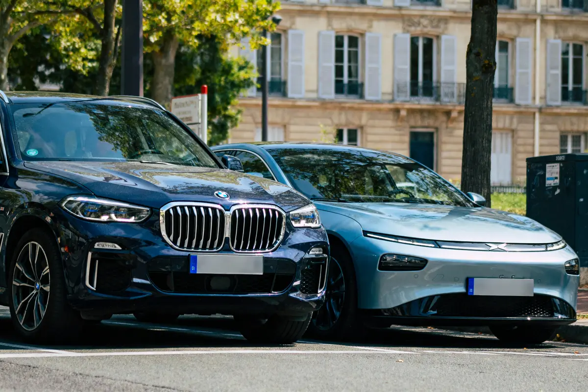 Deux voitures garées côte à côte, devant un bâtiment, sous des arbres ensoleillés.