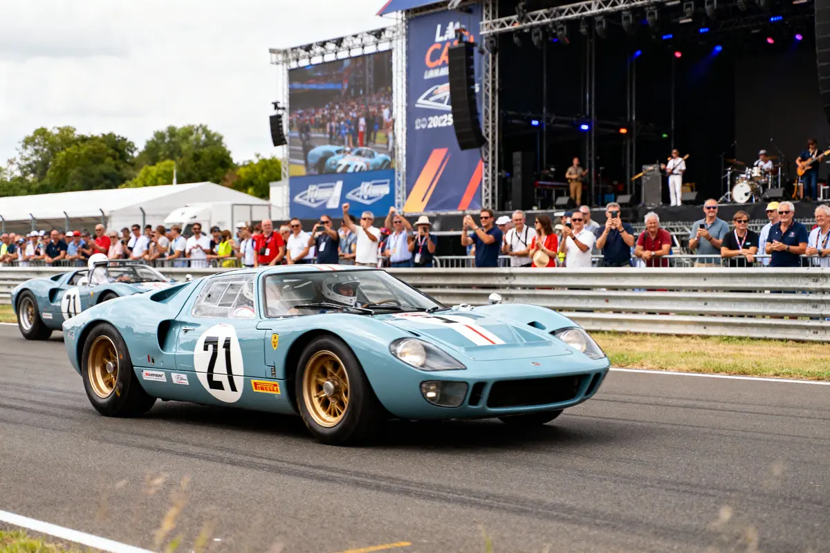Voitures de course anciennes sur circuit, devant spectateurs et scène avec musiciens, ambiance de festival automobile.