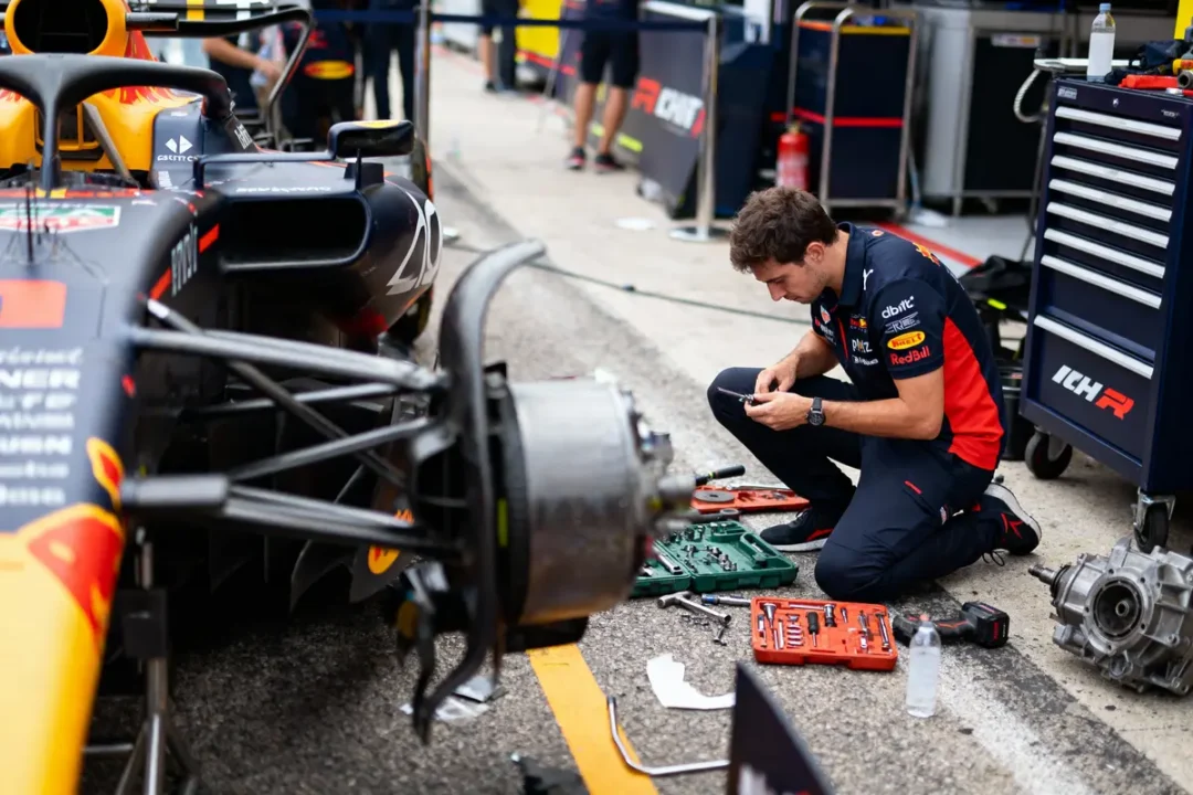 Mécanicien travaillant sur une voiture de course en pit lane, avec outils et pièces autour de lui.