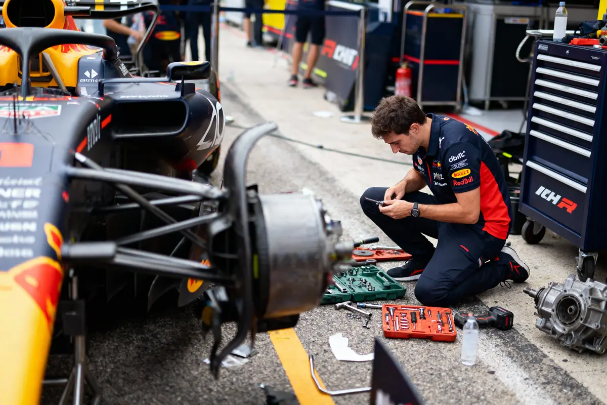 Mécanicien travaillant sur une voiture de course en pit lane, avec outils et pièces autour de lui.