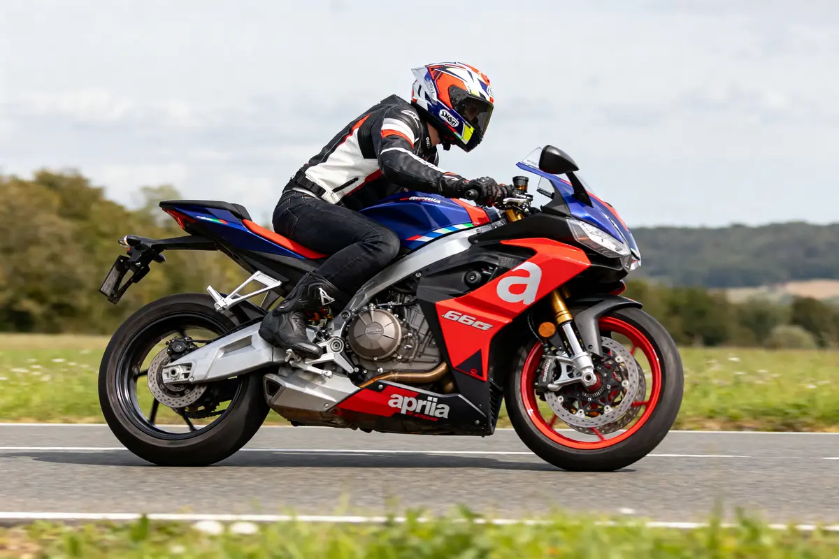 Personne conduit une moto sportive Aprilia rouge et bleu sur une route de campagne, portant équipement de sécurité.