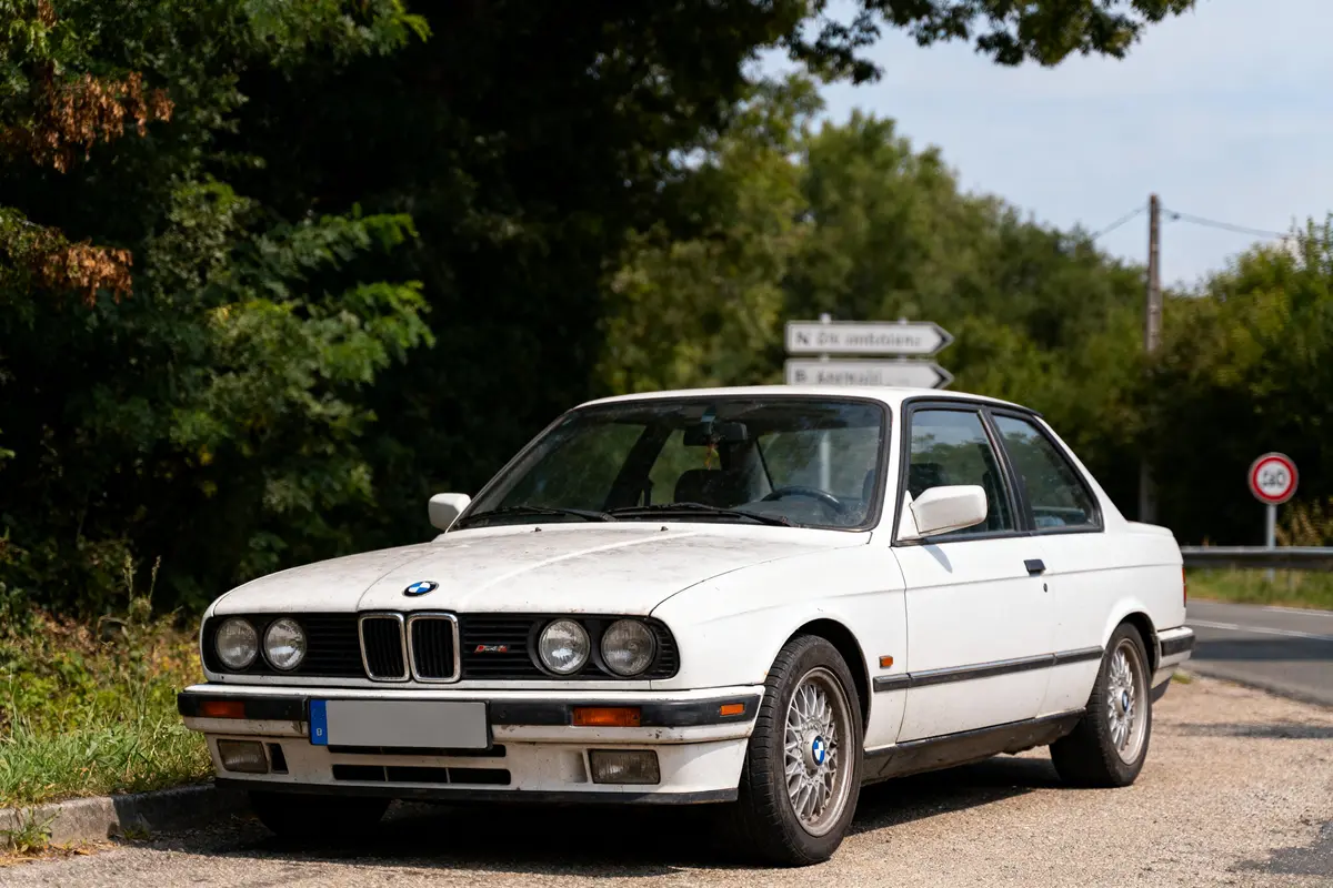 Une voiture BMW blanche ancienne est garée sur le bord d'une route entourée de verdure.