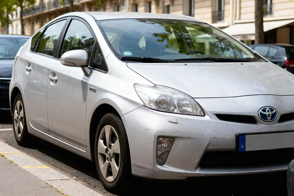 Voiture Toyota Prius argentée garée en ville, rue bordée d'arbres et bâtiments résidentiels.