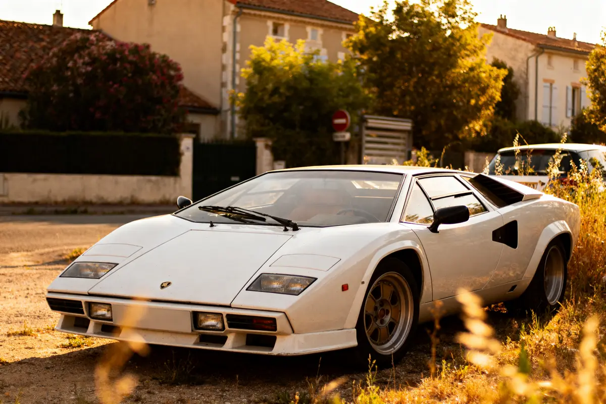 Voiture de sport classique blanche garée, éclairée par le soleil, maisons et végétation en arrière-plan.
