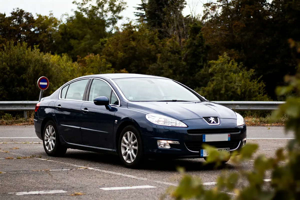 Voiture Peugeot bleue garée sur un parking, arbres en arrière-plan, panneau de signalisation visible.