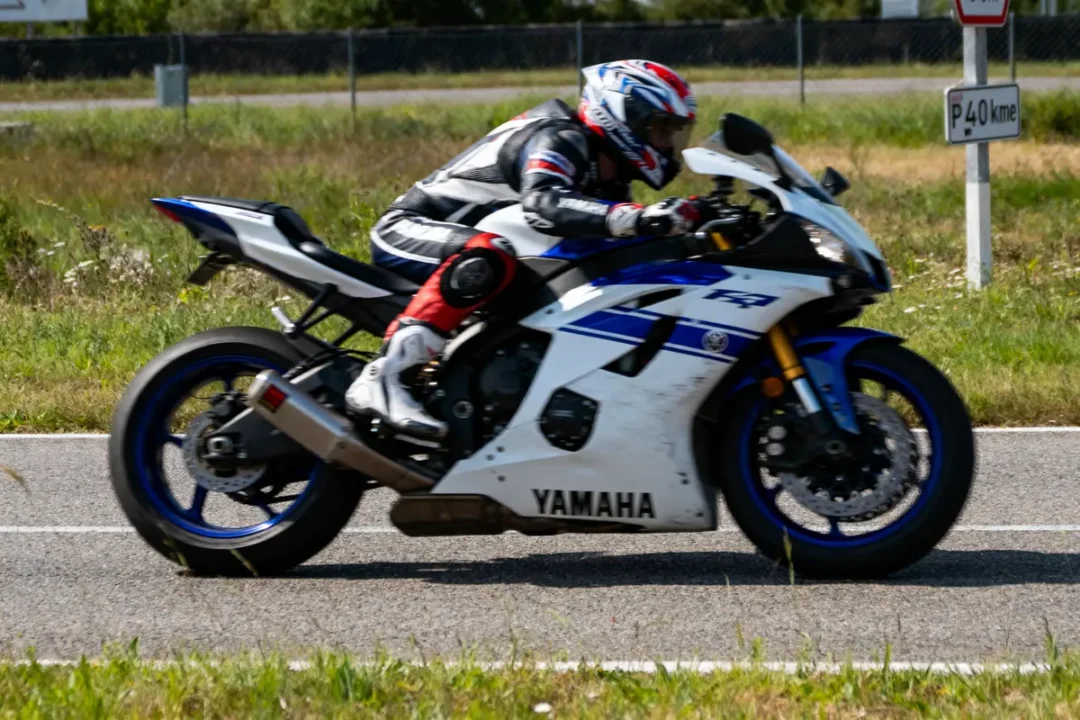Motard en combinaison, sur Yamaha R1, roulant sur circuit. Vitesse indiquée par panneau "P 40 km". Jardin environnant.