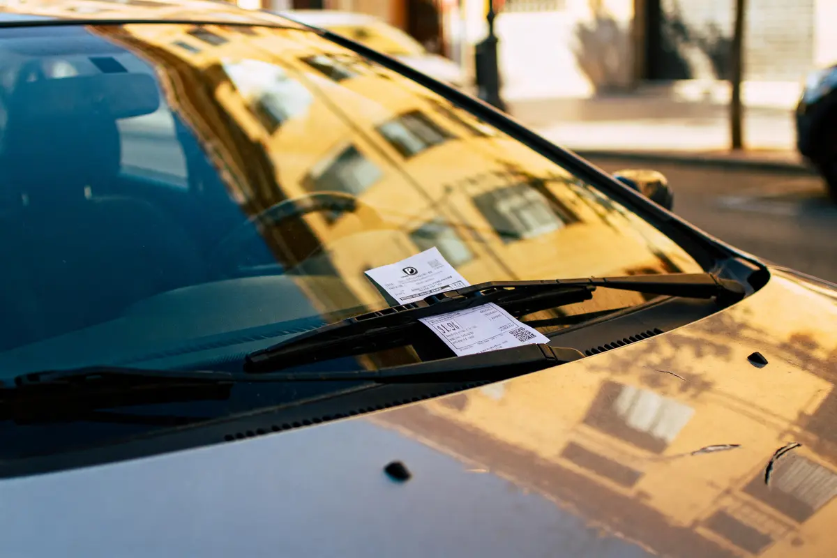 Pare-brise de voiture avec deux tickets stationnement coincés sous essuie-glaces, reflets bâtiments.