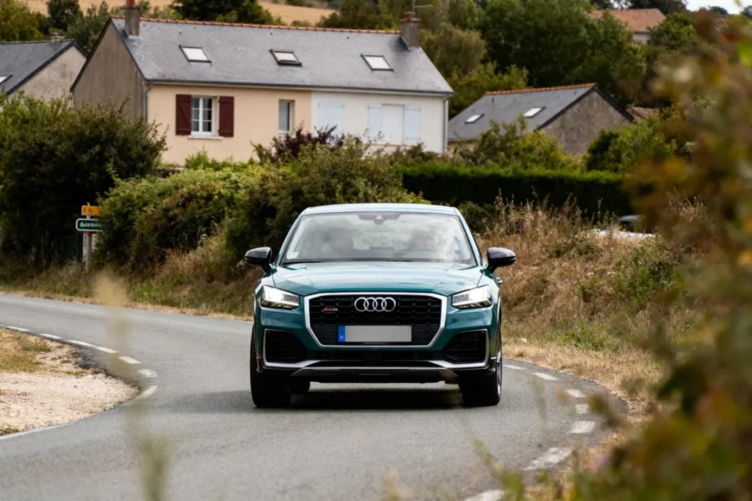 Voiture Audi bleue sur route de campagne, maisons et végétation en arrière-plan, journée lumineuse.
