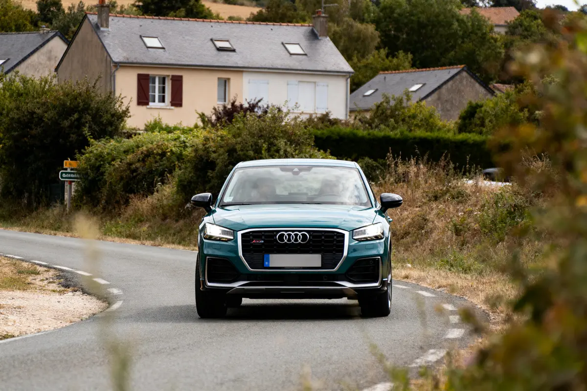 Voiture Audi bleue sur route de campagne, maisons et végétation en arrière-plan, journée lumineuse.