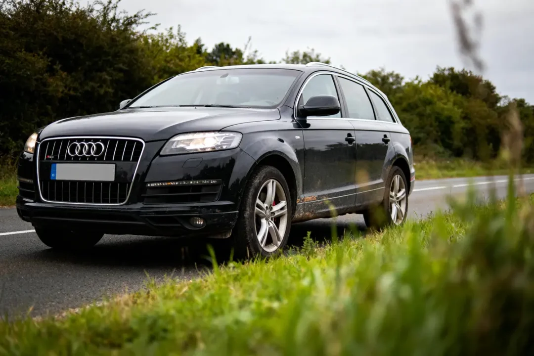 SUV noir Audi Q7 roulant sur une route bordée d'herbe, ciel nuageux, arbres en arrière-plan.