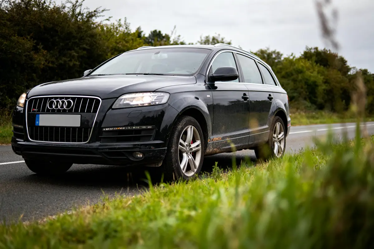 SUV noir Audi Q7 roulant sur une route bordée d'herbe, ciel nuageux, arbres en arrière-plan.