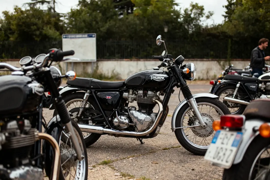 Moto Triumph noire stationnée, entourée d'autres motos, sur un parking extérieur avec arbres en arrière-plan.