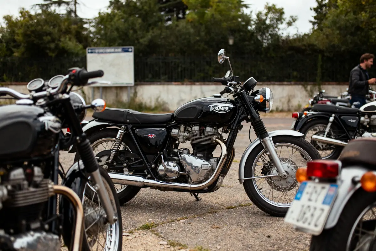 Moto Triumph noire stationnée, entourée d'autres motos, sur un parking extérieur avec arbres en arrière-plan.