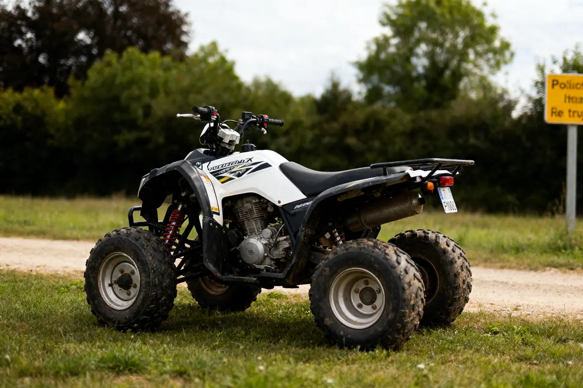 Quad blanc stationné sur herbe, avec arbres et panneau jaune en arrière-plan.