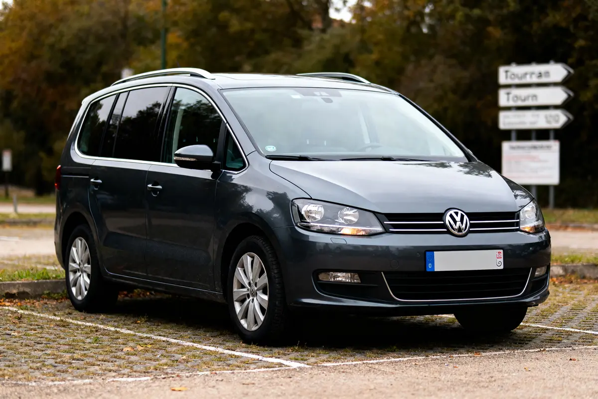 Monospace gris Volkswagen stationné, fond flou avec panneaux routiers et arbres, vue trois quarts avant.