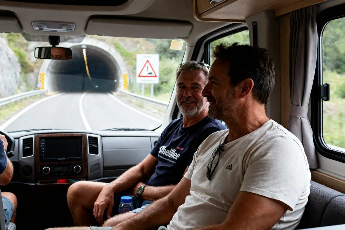 Deux hommes souriants dans un camping-car, roulant vers un tunnel. Paysage verdoyant visible à l'extérieur.