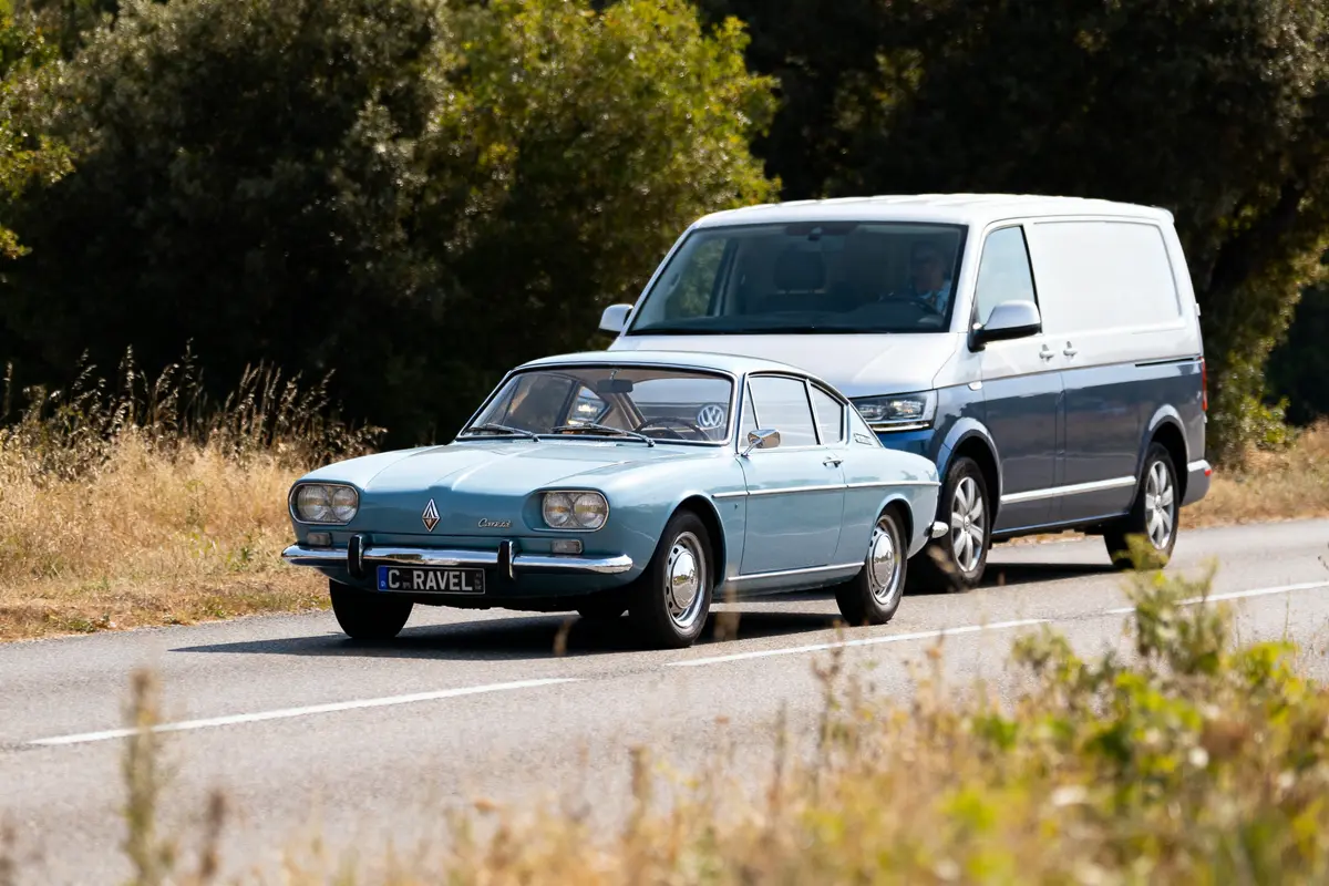 Voiture ancienne bleue suivie d'une camionnette blanche sur une route bordée de verdure.