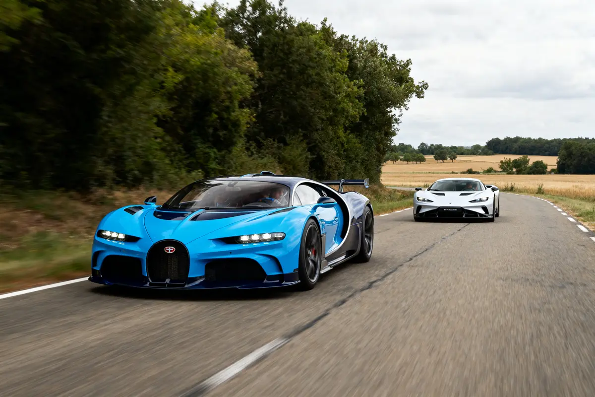 Deux voitures de sport, bleue et blanche, roulant sur une route de campagne bordée d'arbres.